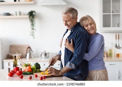 Happy Wife Attractive Senior Lady Hugging Her Husband Chef Cooking Healthy Delicious Dinner For Family, Loving Man Taking Care Of His Partner, Side View, Copy Space, White Kitchen Interior