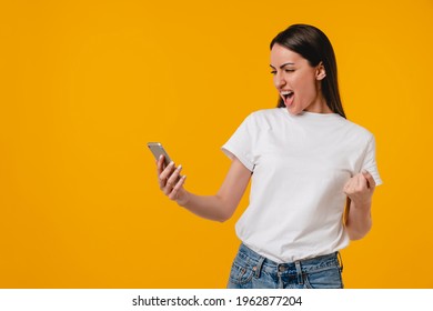 Happy As A Winner Excited Young Hispanic Latin-american Woman Using Smart Phone Isolated Over Yellow Background. Emotional Young Caucasian Female Won The Lottery Big Sum Of Money, Huge Success