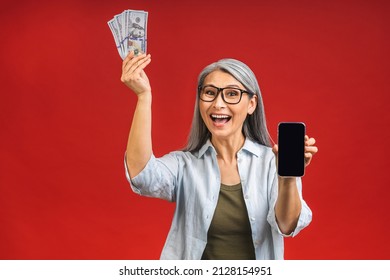 Happy Winner! Image Of Mature Aged Happy Asian Senior Woman Standing Isolated Over Red Background Wall  Holding Money Using Mobile Phone. Showing Phone Screen.