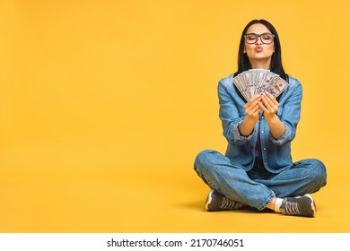 Happy winner! Portrait of a cheerful young woman holding money banknotes and celebrating victory isolated over yellow background. Sitting on the floor in lotus pose.