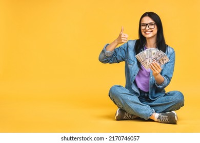 Happy winner! Portrait of a cheerful young woman holding money banknotes and celebrating victory isolated over yellow background. Sitting on the floor in lotus pose.
