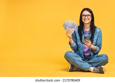 Happy winner! Portrait of a cheerful young woman holding money banknotes and celebrating victory isolated over yellow background. Sitting on the floor in lotus pose. Using mobile phone.