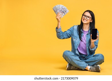 Happy winner! Portrait of a smiling cheerful woman showing blank smartphone isolated over yellow background. Sitting on the floor in lotus pose, holding money bills. 