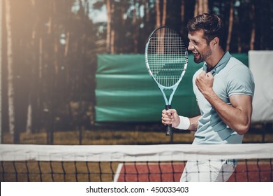 Happy winner. Side view of happy young man in polo shirt holding tennis racket and gesturing while standing on tennis court - Powered by Shutterstock