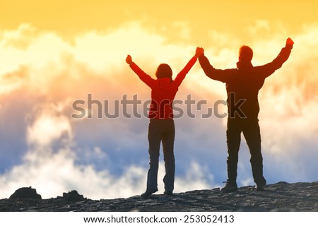 Happy winners reaching life goal - success people at summit. Business achievement concept. Two person couple together arms up in the air of happiness with accomplishment in the clouds at sunset.