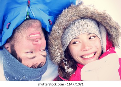 Happy Winter Couple. Cropped View Of The Faces Of A Joyful Young Interracial Asian / Caucasian Couple Lying On Their Backs In Snow With Their Heads Close Together.