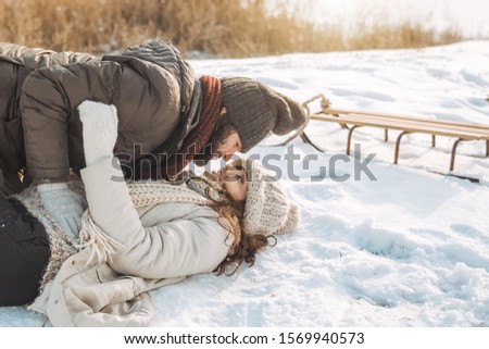 Happy winter couple smiling having fun, fooling around like children in winter park near a snowy river. Young beautiful woman and handsome man in winter warm casual clothes laying on snow playing in