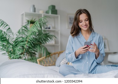 Happy Woman With Appealing Look, Holds Modern Cell Phone, Downloads Song To Playlist, Listens Music With Earphones, Sits Crossed Legs On Comfortable Bed, Has Charming Smile On Face, Uses Free Wifi