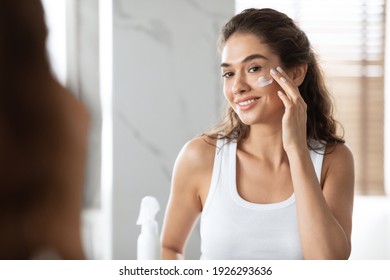 Happy Woman Applying Face Cream Moisturizing And Caring For Skin Smiling To Her Reflection In Mirror Standing In Modern Bathroom At Home. Facial Skincare And Pampering. Selective Focus - Shutterstock ID 1926293636