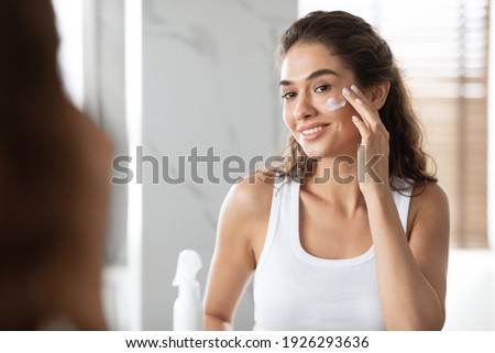 Happy Woman Applying Face Cream Moisturizing And Caring For Skin Smiling To Her Reflection In Mirror Standing In Modern Bathroom At Home. Facial Skincare And Pampering. Selective Focus