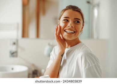 Happy woman applying hydrating moisturizer on her face sitting in bathroom. Home beauty routine - Powered by Shutterstock