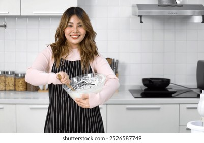 A happy woman in an apron, mixing batter in a clear glass bowl with a wooden spatula. Standing in a clean and modern kitchen - Powered by Shutterstock