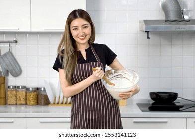 A happy woman in an apron, mixing batter in a clear glass bowl with a wooden spatula. Standing in a clean and modern kitchen - Powered by Shutterstock