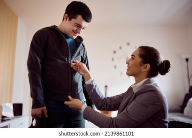 Happy Woman Assisting Man With Down Syndrome In Getting Dressed And Zipping His Jacket. 