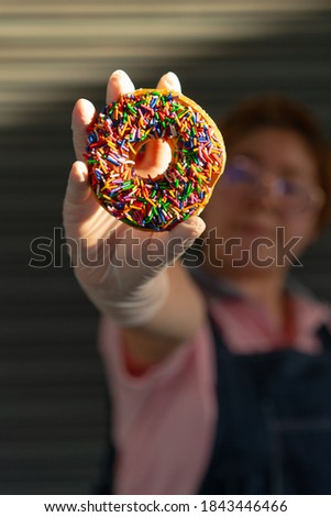 Happy woman baker wearing apron selling chocolate frosted donut sprinkles with a hand wearing latex gloves with her smile. Playful and joyful tasty sugary comfort food for customers. Focus on donut.