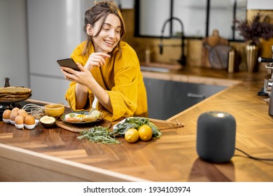 Happy woman in bathrobe controlling home devices with a voice commands, speaking to a smart column during breakfast time on the kitchen at home. Smart home concept - Powered by Shutterstock