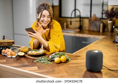 Happy woman in bathrobe controlling home devices with a voice commands, speaking to a smart column during breakfast time on the kitchen at home. Smart home concept