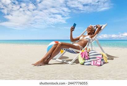 Happy woman at the beach on beach deck chair, sunbathing, uses mobile phone, in a sunny day with blue sky, concept a summer beach holiday, online shopping, booking travel, and resorts accommodations - Powered by Shutterstock