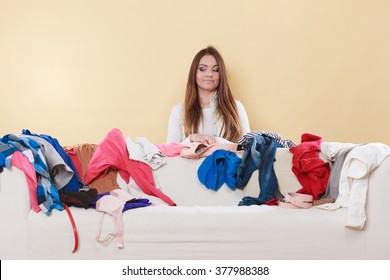 Happy Woman Behind Sofa Couch In Messy Living Room. Young Girl Surrounded By Many Stack Of Clothes. Disorder And Mess At Home.