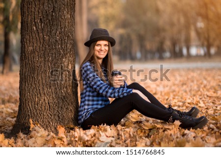 Happy woman in black hat drinking hot coffee in park under fall foliage. Beautiful young modern smiling woman on coffee break . Fall concept