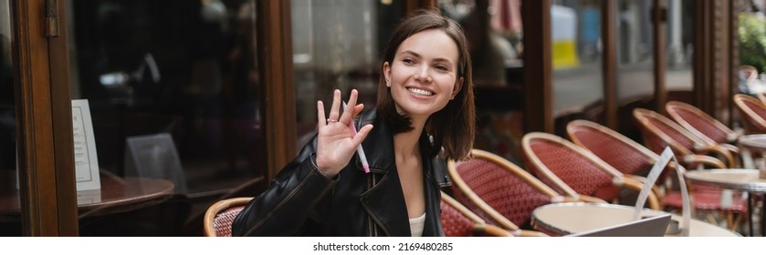 Happy Woman In Black Jacket Holding Pen And Waving Hand In French Outdoor Cafe, Banner