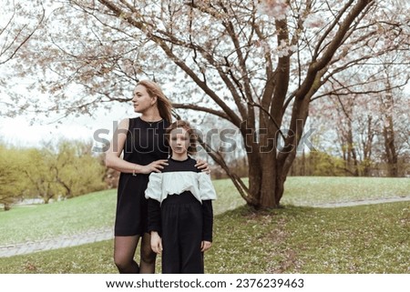 Happy woman with blond hair hugging girl in black blouse while standing under tree in park