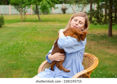 Happy Woman In Blue Dress Holding Ginger Imitation Cat Outside, Outdoors In Summer In Park Sitting In Armchair On Lawn Stroking Her Beloved Pet Holding Him In Her Arms