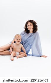 Happy Woman In Blue Dress Looking At Camera Near Infant Kid Sitting On White Background