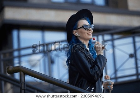 Happy woman with blue hair enjoying the music. Portrait of a diverse female person wearing wireless headphones and licking a lollipop candy in the city street
