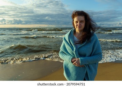 Happy Woman In A Blue Knitted Poncho On The Background Of Sea Waves, With Hair Developing In The Wind. Mature Woman Posing On The Beach In Autumn.