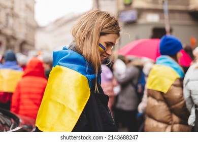 Happy woman with blue and yellow Ukrainian flag on her cheek  wrapped in Ukrainian flag at a demonstration. Support Ukraine and woman with Ukraine Flag.