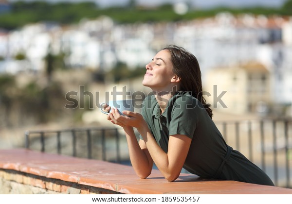 Happy woman breathing fresh air enjoying sun and drinking coffee from balcony in a beach town