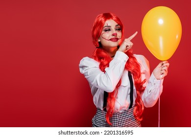 Happy Woman With Bright Hair And Clown Makeup Pointing At Yellow Balloon Isolated On Red