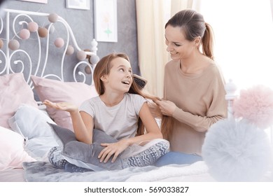 Happy woman brushing her daughter's hair, sitting on bed - Powered by Shutterstock