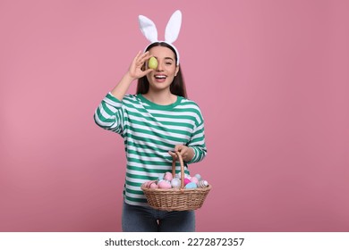Happy woman in bunny ears headband holding wicker basket of painted Easter eggs on pink background