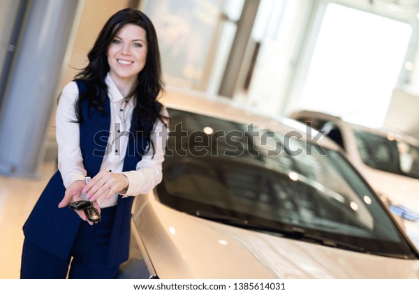 Happy woman buyer possing with keys near her\
new vehicle in car\
dealership