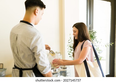 Happy Woman Buying Organic Flour On Her Own Container At A Sustainable Grocery Store While Talking To A Latin Worker