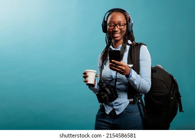 Happy Woman With Camera Device Enjoying Songs While Browsing Internet On Phone. Joyful Professional Photographer With Wireless Headphones And Modern Smartphone Listening To Music On Blue Background.