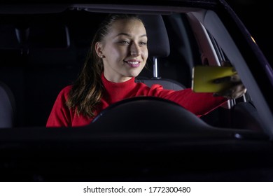 Happy Woman In Car At Night Stopped Parked And Talking With Family By Video Call. Smiling Female Driver Sitting Inside Car Makes Selfie. Concept: Car Blog, Car Travel Broadcast, Automobile Vlogger