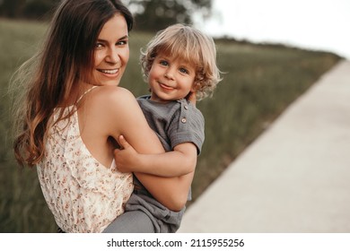 Happy woman carrying cute blond boy and looking at camera with smile on blurred background of path and lawn in park