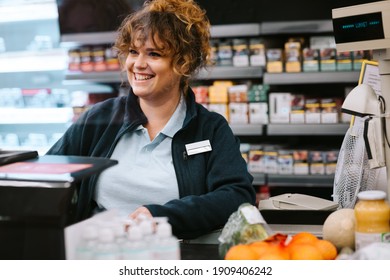 Happy Woman Cashier At A Supermarket Attending Customer. Woman Working At Grocery Store Checkout Talking With Customer And Smiling.