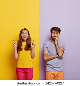 Happy Woman In Casual Clothes, Keeps Fingers Crossed, Believes In Good Luck, Sad Dejected Man Feels Bored, Stands Near, Pose Against Yellow And Purple Striped Background. Body Language Concept