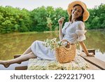 a happy woman with a chamomile flower in her hands sits on a pier by the lake and looks at the camera smiling
