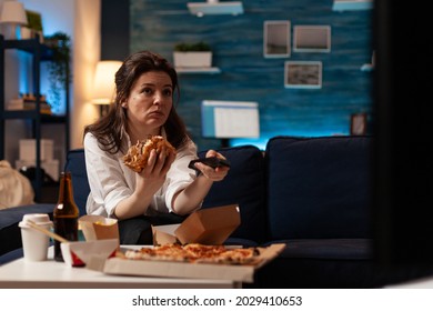 Happy woman changing channels using remote watching entertainment movie on television eating tasty delicious burger in evening. Caucasian female enjoying takeaway food home delivered