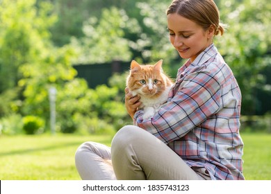 Happy woman in checked shirt hugging and embracing with tenderness and love domestic ginger cat, sitting on grass outdoors in sunny day. Love to the animals