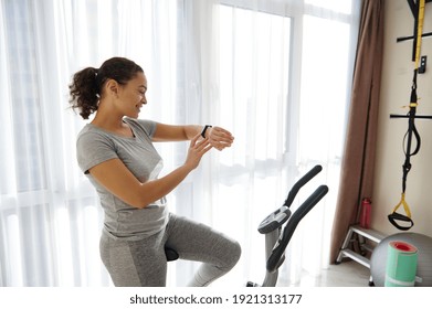 Happy Woman Checking Her Fitness Tracker And Heart Rate While Standing On A Spin Bike After Cardio Workout. Fit Woman Enjoying Workout At Home