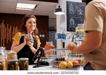 Happy woman at checkout counter in zero waste shop selling healthy lentils in jar to vegan customer with green living lifestyle. Client buys food in local neighborhood store from friendly seller