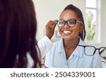 Happy woman chooses new eyewear. Optician offers new design to customer. Sales assistant at store or optometrist at clinic helps smiling young African American woman put on glasses