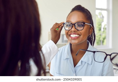 Happy woman chooses new eyewear. Optician offers new design to customer. Sales assistant at store or optometrist at clinic helps smiling young African American woman put on glasses - Powered by Shutterstock