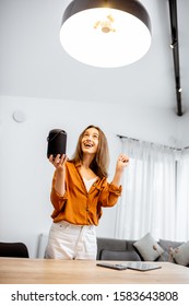 Happy Woman Controlling Home Light With A Smart Speaker In The Living Room. Concept Of A Smart Home And Light Control With Voice Command